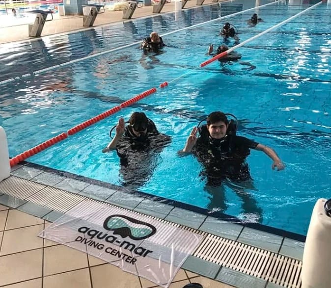 Doi cursanți echipați la suprafața piscinei, lângă bannerul Aqua-Mar